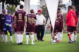 Eine Gruppe von Fußballspielern in kastanienbraunen Trikots und ein Trainer in einem weißen Hemd versammeln sich auf einer Wiese, lächeln und unterhalten sich, einige tragen Beinprothesen, mit einer weißen Flagge im Vordergrund.