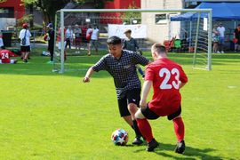 Zwei junge Männer spielen auf einer Wiese Fußball; der eine in einem karierten Hemd spielt den Ball, während der andere in einem roten Trikot mit der Nummer 23 verteidigt. Im Hintergrund sind Zuschauer und ein Tor zu sehen.