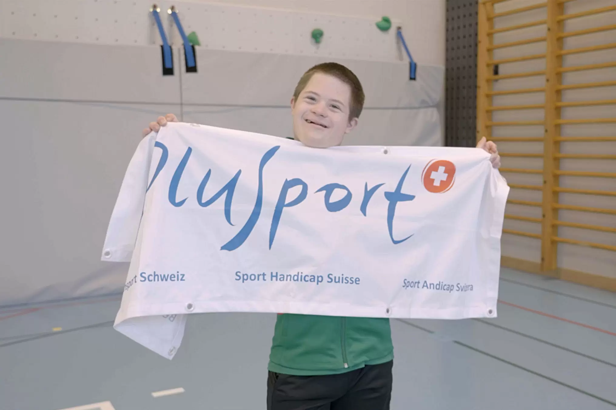 Ein junger PluSport-Teilnehmer hält in einer Turnhalle die PluSport-Flagge in die Luft