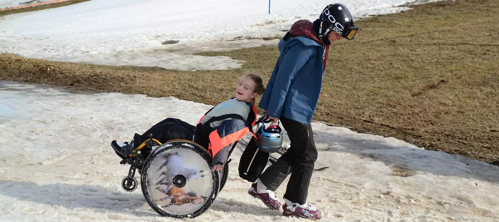 [Translate to Francais:] Eine Teilnehmerin im Rollstuhl wird von der Camps-Leiterin über ein Schneefeld gezogen. Beide lachen.