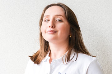 Eine Frau mit langen braunen Haaren und einem weißen Hemd mit Kragen steht vor einer hellen, strukturierten Wand und blickt selbstbewusst in die Kamera.